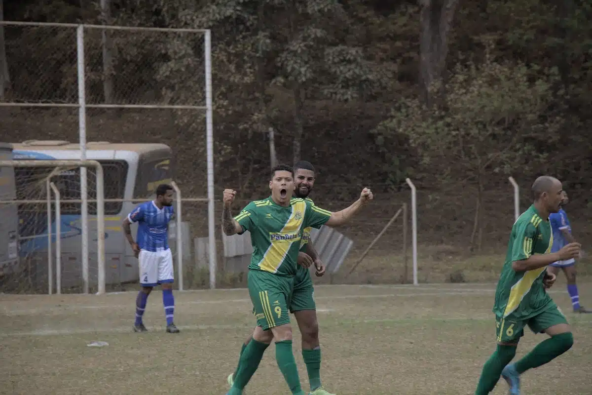 Jogador Kenedy, centro-avante do 8 de Dezembro, ergue os braços e vibra após ter feito o único gol da partida contra o São Caetanense. Atrás dele e à direita da imagem, dois companheiros de equipe, vestindo camisas verdes, com faixa diagonal em amarelo no peito, e calção verde. Ao fundo, zagueiro do São Caetanense, de calção branco e camisa azul.