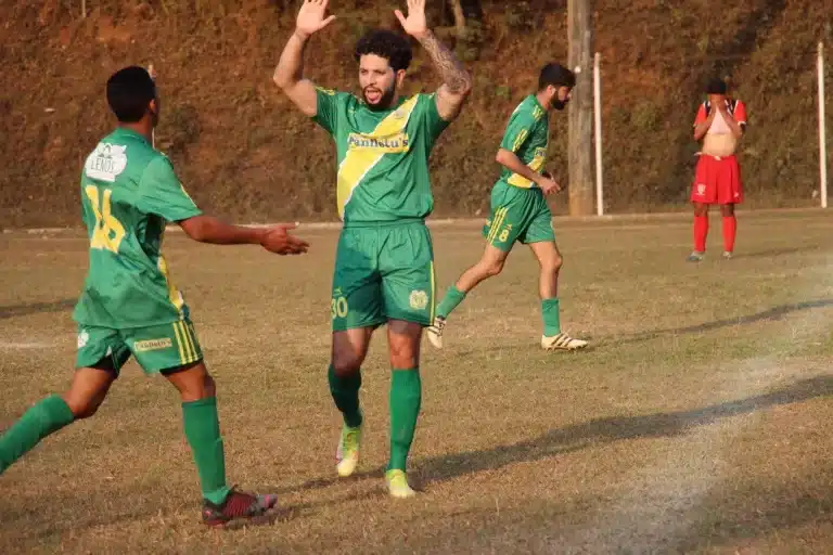Jogador do 8 de Dezembro, com uniforme verde com listra diagonal amarela na camisa, comemora gol junto a um colega, de costas, em partida válida pela 4ª rodada do Campeonato Amador de Mariana