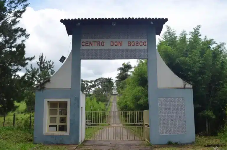 Entrada do terreno, com um arco na cor azul e identificação “Centro Dom Bosco”. Ao fundo da propriedade, depois de um caminho em aclive, cercado por vegetação, vê-se a fachada do prédio do antigo Colégio Dom Bosco