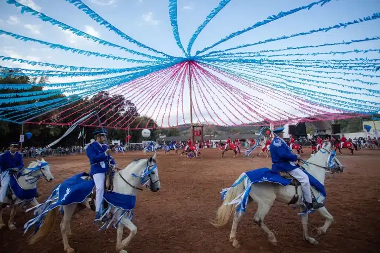 Encenação de batalha entre cristãos e mouros, em Amarantina, distrito de Ouro Preto. Em primeiro plano, cavaleiros com uniformes azuis montados em cavalos brancos. Ao fundo, cavaleiros de uniforme vermelho em cavalos marrons. Um poste, ao centro, suporte fitas amarelas e vermelhas, dispostas radialmente em um círculo