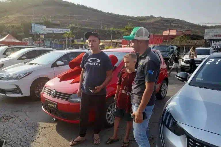 Homem e seu filho, junto com um vendedor, diante de um carro vermelho adquirido no 1º Feirão de Automóveis de Mariana