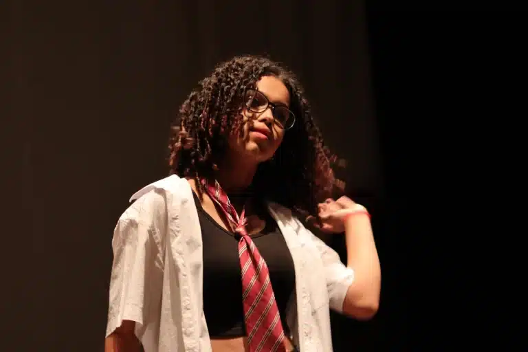 Dançarina apresentando-se na noite de abertura da 6ª edição do Festeco, de cabelos cacheados, usando óculos, camisa branca aberta, gravata vermelha com listras e corpete preto