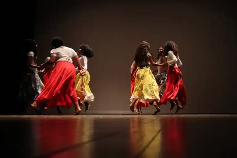 Dançarinas, com roupas coloridas, formam duas rodas no palco, cada uma com 4 integrantes, durante apresentação na noite de abertura do Festeco