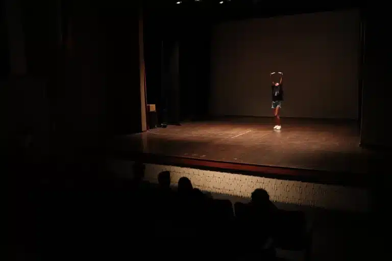 Dançarina, usando tênis branco, short jeans e blusa preta, com os braços levantados, faz apresentação solo no centro do palco, na noite de abertura do Festeco