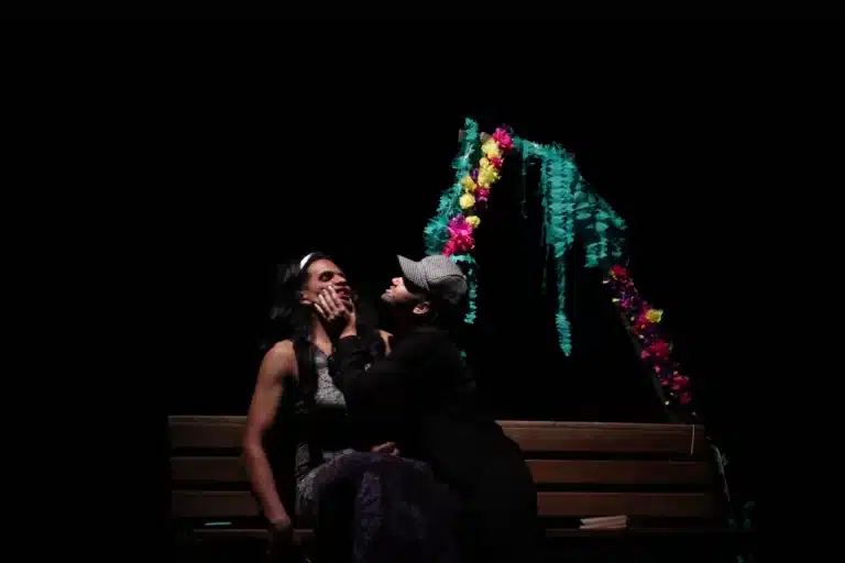 Cena da apresentação de "Muito barulho por nada", na abertura da 6ª edição do Festeco. Assentado no banco, colocado diante de um enfeite colorido, homem de boné e roupas escuras, segura com força o rosto de uma mulher