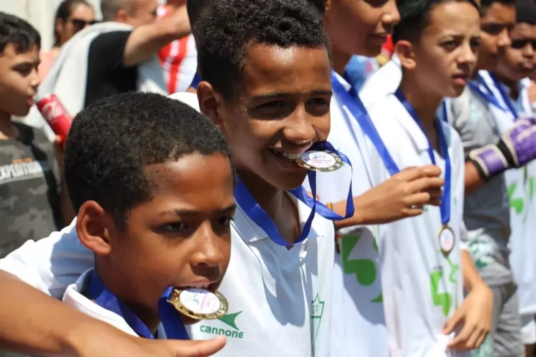Dois jogadores da categoria sub-13, com camisas brancas, seguram as medalhas com os dentes