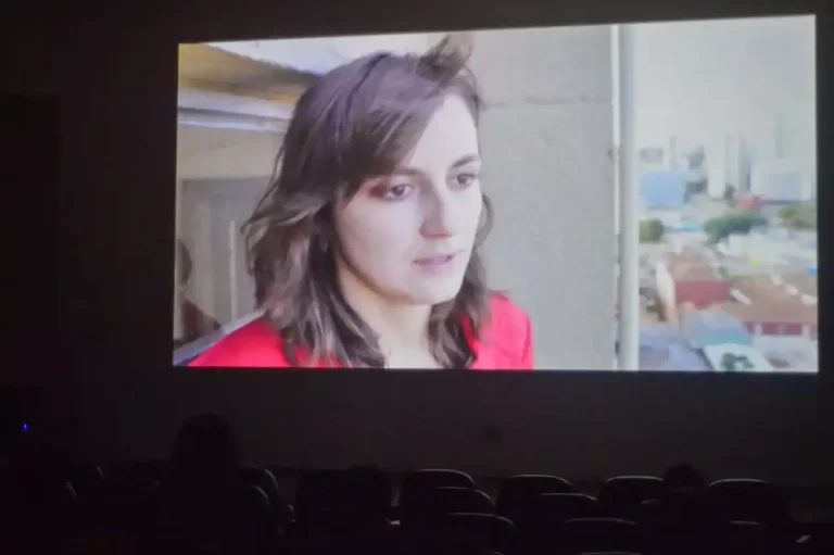 Telão com exibição de um vídeo protagonizado por uma mulher. Abaixo, cadeiras com telespectadores da mostra de cultura latino-americana