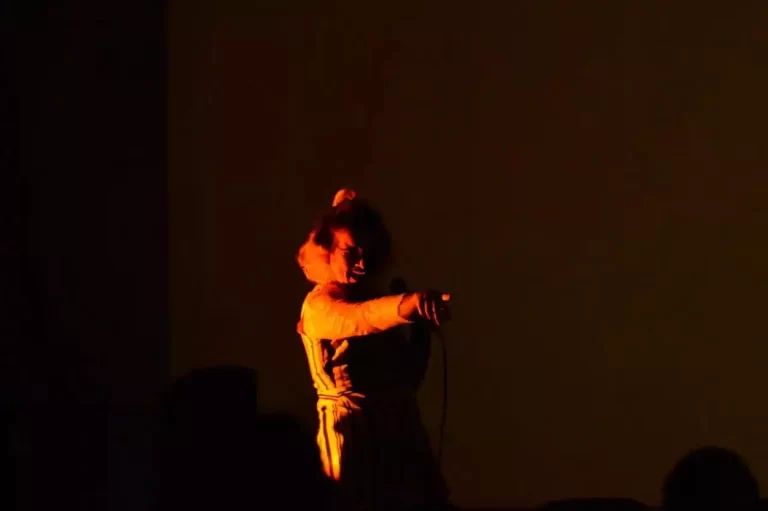 Artista Guga Medeiras no palco, segurando um microfone enquanto apresenta sua performance musical, na mostra de cultura latino-americana, no auditório do anexo do Museu da Inconfidência