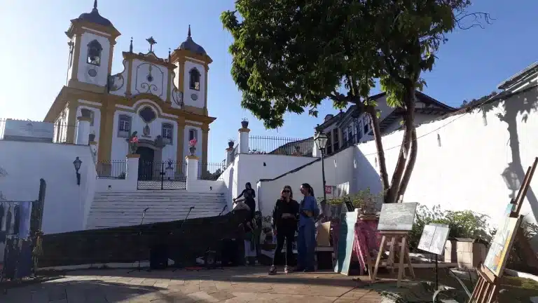 Ao fundo, Igreja Nossa Senhora da Conceição, em Ouro Preto. Em primeiro plano, a praça em frente à Igreja, com exposição de quadros e roupas para venda, e pessoas conversando