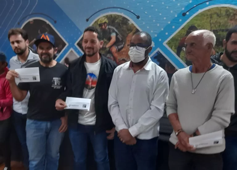 Atletas posam para foto, exibindo papel referente à isenção da taxa de inscrição no Iron Biker, á frente de painel alusivo ao esporte