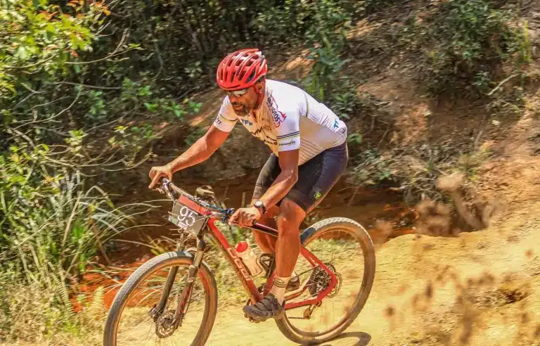 De bermuda branca, capacete vermelho e camiseta branca, com algumas inscrições colorida, Renato Estevão pedala em trilha de terra, tendo ao fundo um barranco com vegetação, competindo na Copa das Vertentes