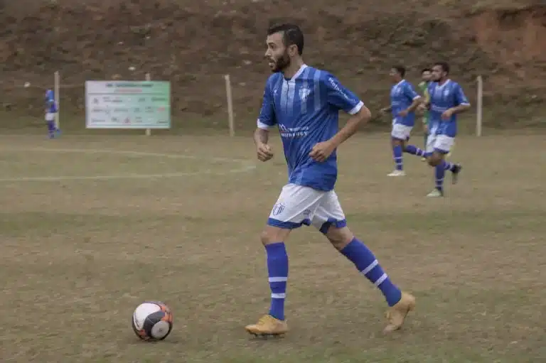 Jogador do São Caetanense, de calção branco e camisa azul, conduz a bola durante uma partida do Campeonato de Futebol Amador de Mariana