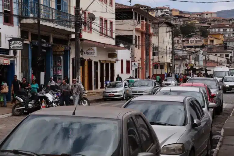 Trânsito na Avenida Salvador Furtado, com uma longa fileira de carros estacionados, com algumas pessoas na calçada, ao fundo, do outro lado da rua