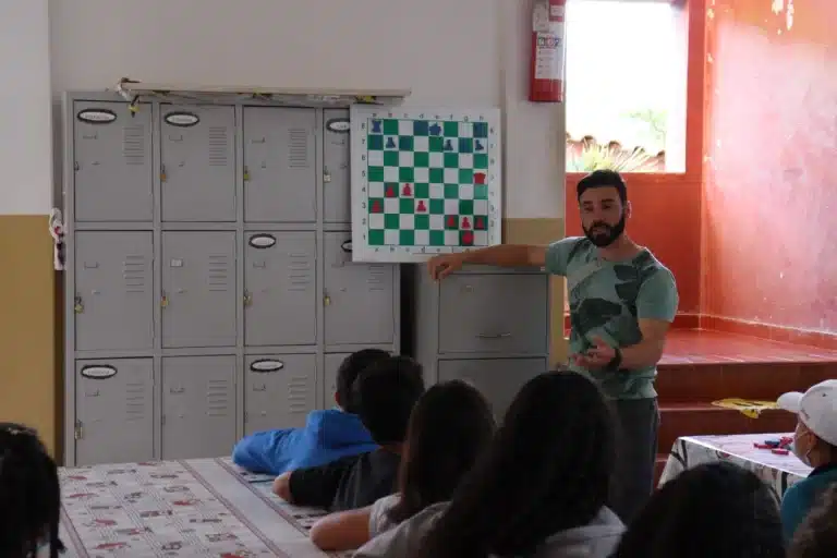 Miguel Magalhães, ao lado de um tabuleiro de xadrez, colocado em uma parede,com casas em verde e branco, explicando aspectos táticos do jogo