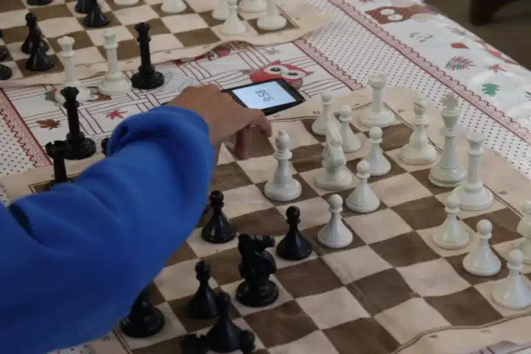 Tabuleiro de xadres, com peças em branco e preto, durante execução de um lance de partida simultânea, executado por um aluno do qual vê-se apenas o braço direito, com roupa azul. Ao fundo, um celular utilizado para a contagem do tempo de jogada de cada enxadrista.