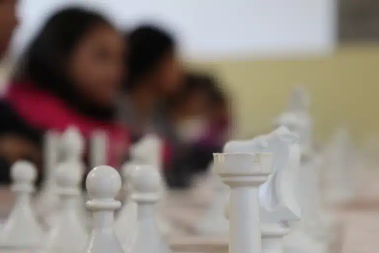 Em primeiro plano, peças brancas do jogo de xadrez, tendo ao fundo a silhueta de integrantes do Clube de Xadrez
