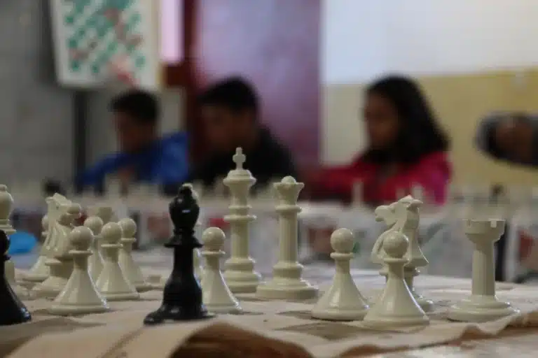 Em primeiro plano, peças brancas e uma preta sobre tabuleiro de xadrez, tendo ao fundo as silhuetas de três integrantes do Clube de Xadrez