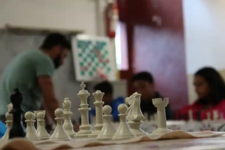 Em primeiro plano, peças brancas do jogo de xadrez. Mais atrás, o professor Miguel executa jogada na partida simultânea,. Vê-se também , as silhuetas de dois integrantes do Clube de Xadrez e, mais ao fundo, um tabuleiro, pendurado na parede, usado para as explicações táticas do jogo