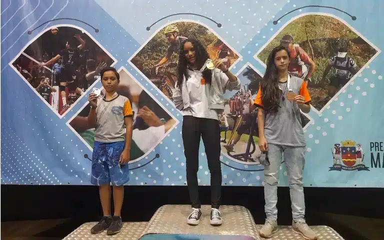 No pódio, colocado à frente de painel dos Jogos Escolares de Mariana, Luan, Claudilene e Bruna mostram as medalhas conquistadas no Módulo 1 de Xadrez