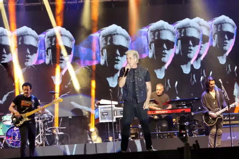No fundo do palco dos shows de abertura da Expo Mariana, um grande painel exibe várias imagens de Rogério Flausino, postado no centro do palco, completamente vestido de pretorio Flaus