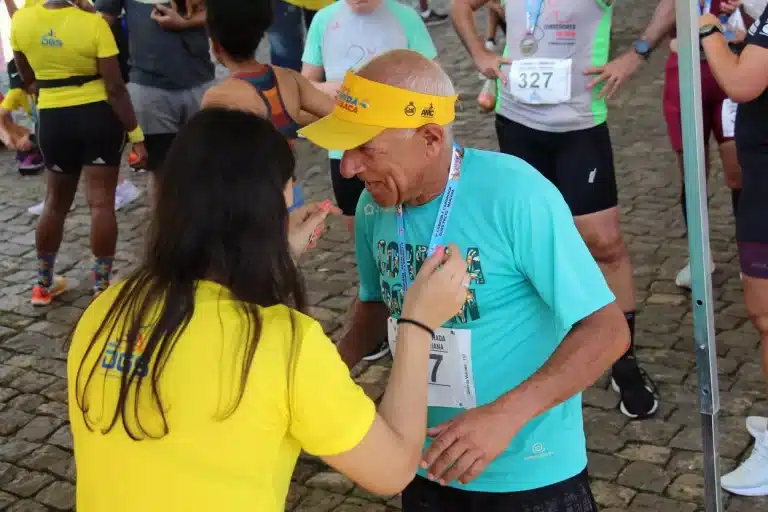 ao término da prova, senhor idoso, usando camiseta azul e short branco, recebe sua medalha de participação na 9ª Caminhada Ouro Preto - Mariana, das mãos de integrante da equipe de organização, usando camiseta amarela, com a inscrição "DGS Sports" às costas.