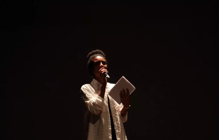 EStudante vestida de branco, com microfone e caderno nas mãos, declama poemas durante abertura do Festival de Teatro Comunitário
