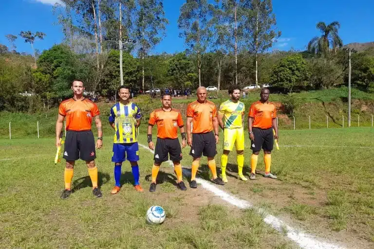 Quatro árbitros, de calções pretos e camisas alaranjadas com detalhes em preto, posam para foto oficial, junto com os capitães das equipes que disputaram a primeira partida da final da 1ª Divisão
