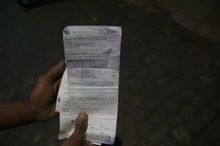 À frente de um fundo escuro, duas mãos seguram contas de água da Saneouro, com valores considerados abusivos