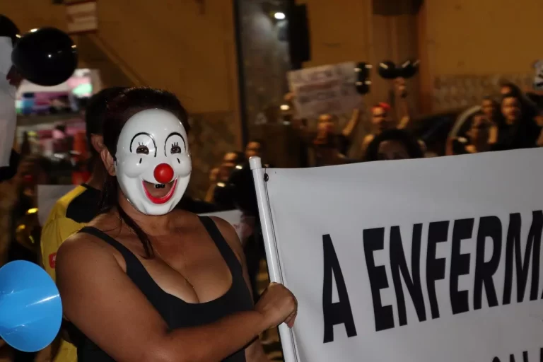 Manifestante, na passeata dos profissionais de Enfermagem, usando máscara branca, com nariz vermelho, segura faixa de protesto