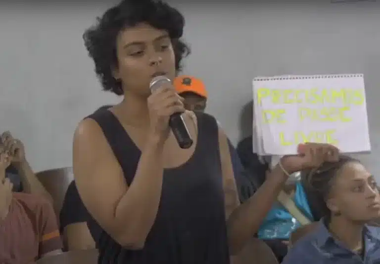 Estudante, de blusa preta, segurando um cartaz feito em caderno,fala ao microfone durante Audiência Pública da Câmara de Ouro Preto, que discutiu a implantação do passe livre estudantil no município.