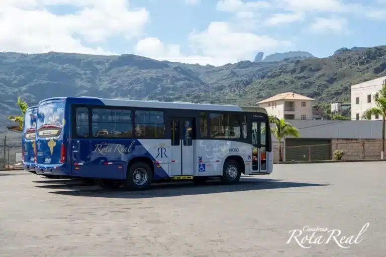 Ônibus, com pintura em azul e branco, do Cosórcio Rota Real, em rua de Ouro Preto, vendo-se ao fundo o Pico do Itacolomi
