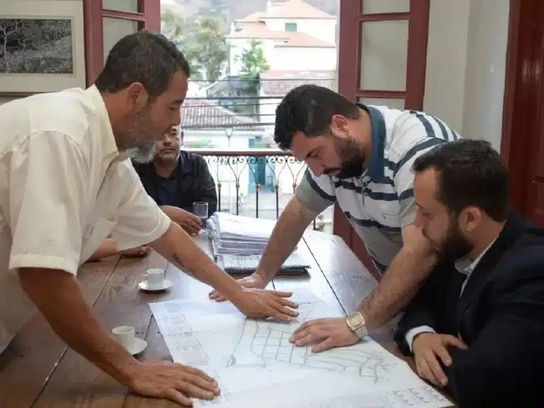 Homens, em torno de uma mesa, dois em pé e dois assentados, discutem detalhes do Diagnóstico pluviométrico e de esgotamento sanitário do Residencial Dom Bosco