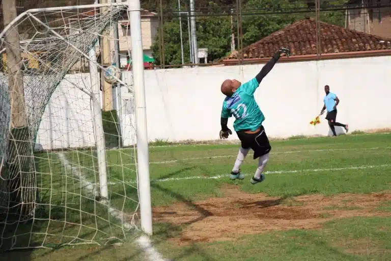 Goleiro do Olimpic, com calção preto e camisa azul, salta mas não consegue interceptar a bola na falta cobrada por jogador do 08 de Dezembro