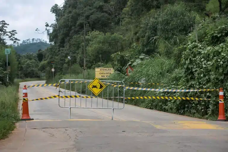 Trecho de estrada com interdição formada por cones, fitas, grade e placa, indicando risco de desmoronamentos no período chuvoso