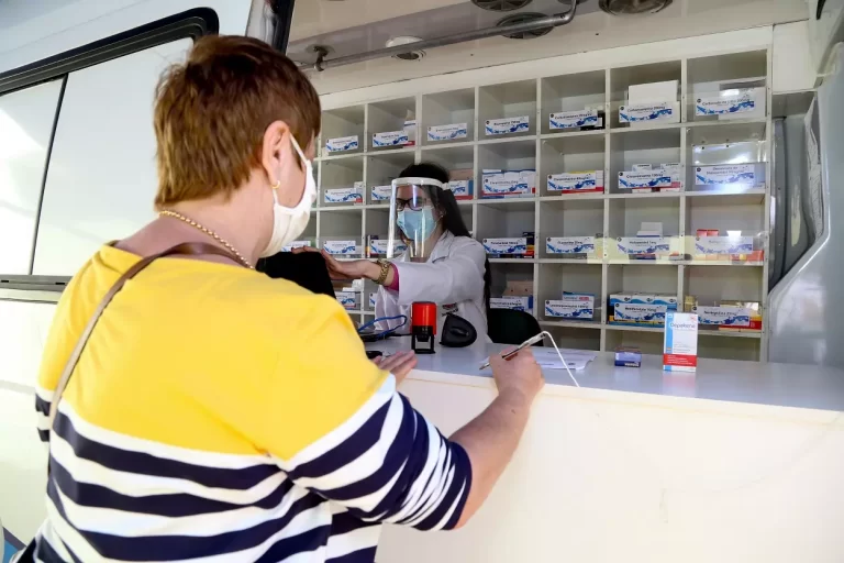 Mulher com máscara e blusa amarela, com listras azuis e brancas, assina documento de recebimento de medicamentos da Farmácia Móvel. Ao fundo, atendente de máscara, diante de prateleira de medicamentos dentro do veículo