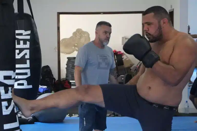 Atleta, sem camisa e luvas de boxe, executa chute em saco de treino, observado pelo treinador de MMA, André Benkei