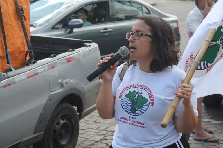 Letícia Oliveira, coordenadora do MAB, com blusa branca e símbolo do movimento no peito, usando óculos, usa o microfone durante a Marcha dos Atingidos