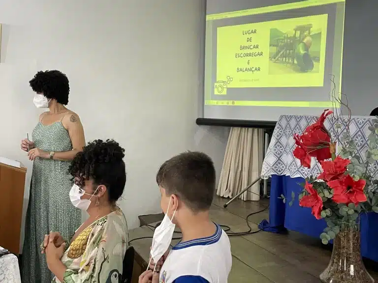 Diante de uma tela exibindo fotografia de um parquinho infantil, com os dizeres "Lugar de brincar escorregar e balançar", aluno se apresenta na abertura da exposição de poesias e fotografias