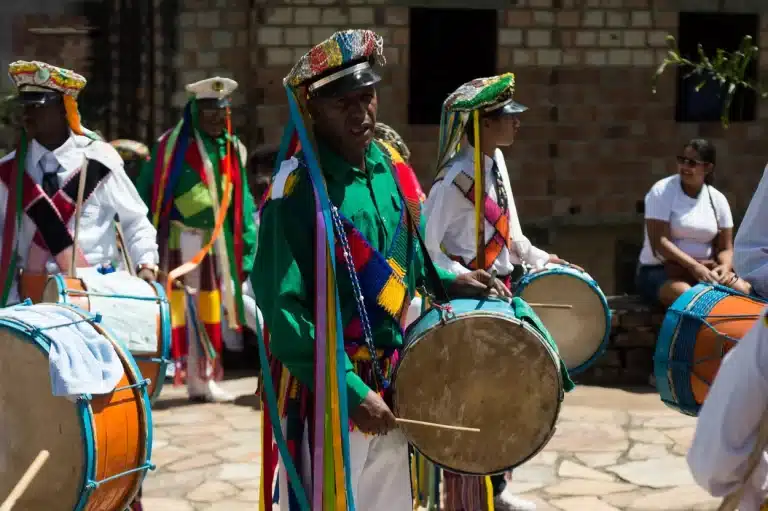 Quatro homens aparamentados, dois deles em roupas brancas e dois com calças brancas e verdes, todos com bonés de aba, dos quais pendem fitas coloridas, tocam tambores em cortejo da Festa do Reinado