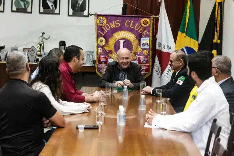 MPessoas em torno de uma mesa, com bandeiras ao fundo, durante assinatura do convênio para atendimento a moradores de Mariana no Hospital dos Olhos de Ouro Preto