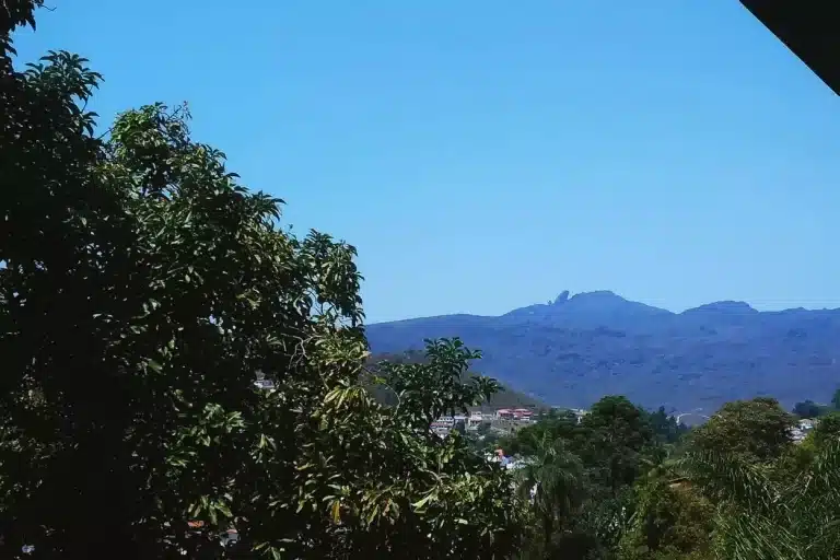 Pico do Itacolomi, visto de uma janela, sob sol azul, com vegetação em primeiro plano