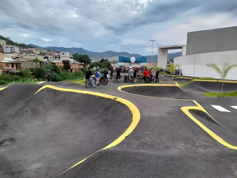 Pessoas em bicicletas, com pórtico azul ao fundo, assistem à cerimônia de inauguração da pista de pump track construída em Mariana.