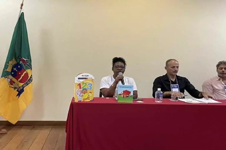 SEntada atrás de uma mesa coberta por toalha vermelha, tendo à sua esquerda dos vereadores e à direita a bandeira de Mariana, Cristina Pereira, com o microfone na mão, faz pronunciamento na VI Conferência Municipal dos Direitos da Criança e do Adolescente