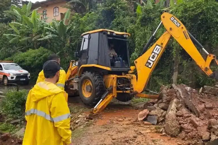 Pessoas trajando capas amarelas observam trabalho de equipamento de remoção de material de desmoronamento