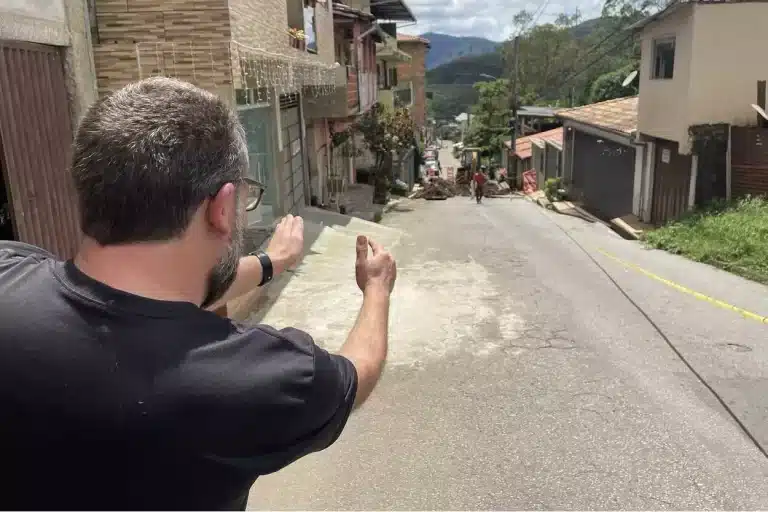 Morador da rua João Batista, de costas, estende os braços apontando os problemas da rua João Batista, em Passagem de mariana, vista do alto para baixo, percebendo-se, ao fundo, a movimentação de máquinas e equipamentos para o reparo da via.
