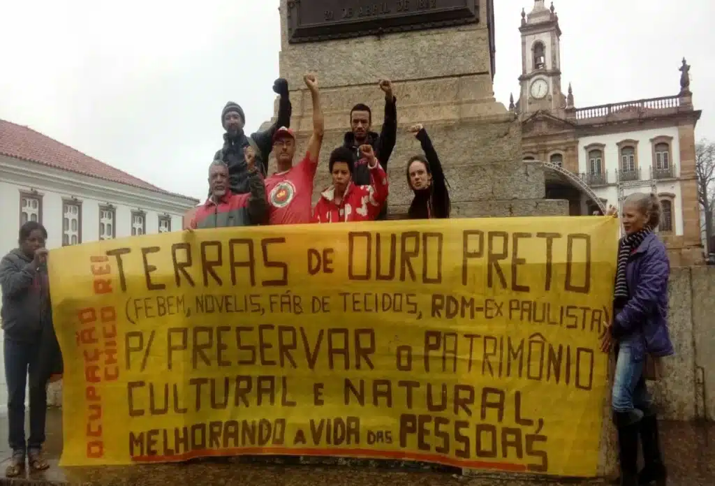 Manifestantes portam faixa, em frente à estátua de Tiradentes, pedindo a destinação de terras para fins sociais
