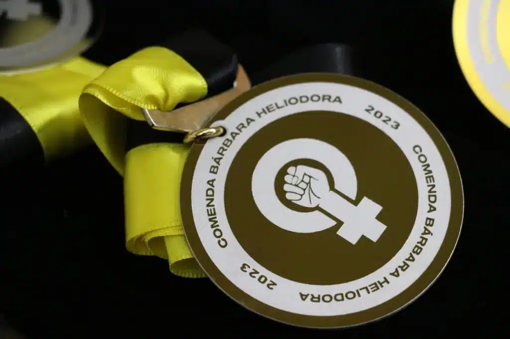 A Comenda Bárbara Heliodora exibe um punho erguido, em sobreposição ao símbolo do gênero feminino, para simbolizar a força, a solidariedade e o apoio às mulheres