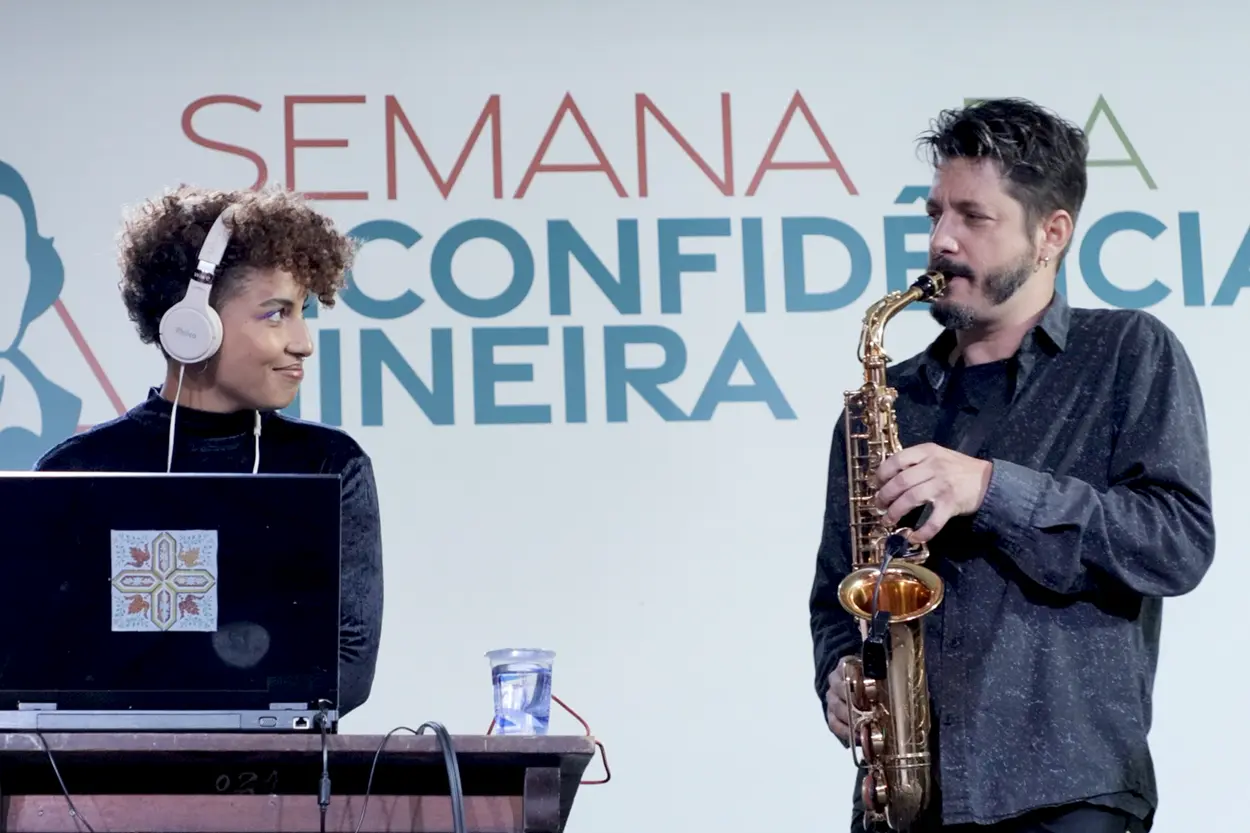 A imagem mostra duas pessoas na frente dos dizeres: Semana da Inconfidência Mineira, do lado esquerdo vemos a DJ Viviane Bascelos, ela está com um fone, em frente a um computador e olha para o Chumbinho que está do lado direito da imagem tocando um saxofone.