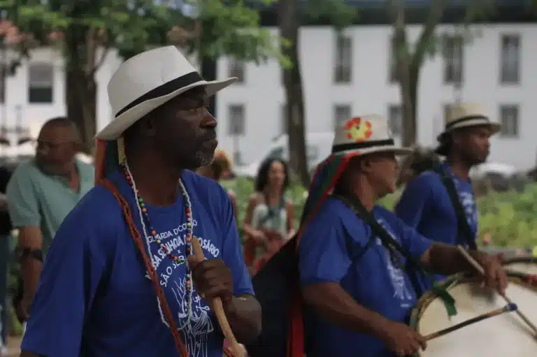 Homens negros com chapéus e camisetas azuis tocam instrumentos em cortejo do congado em Mariana, valorizando a herança africana.