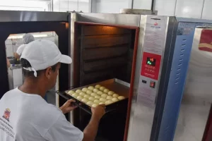 A imagem mostra um homem retirando uma fornada de pães de queijo de um forno durante inauguração da Padaria Escola